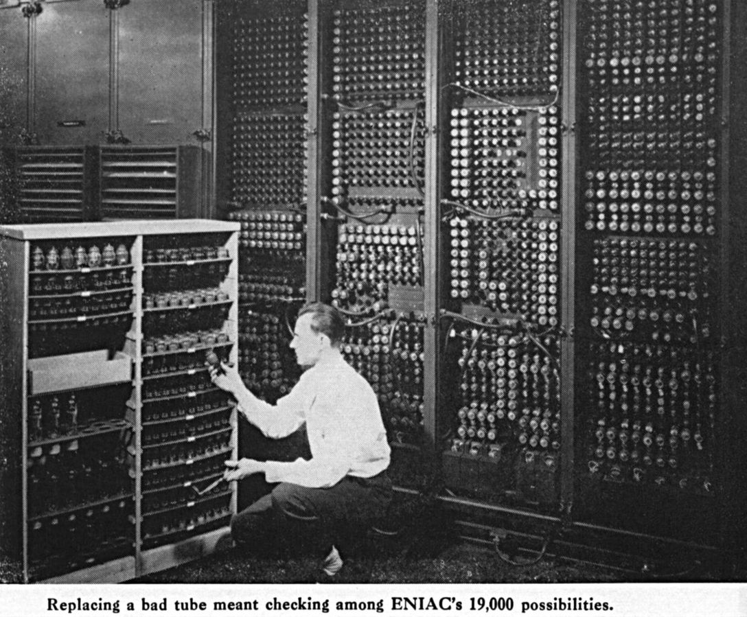 A man kneels in front of the giant early computer ENIAC to change a tube.