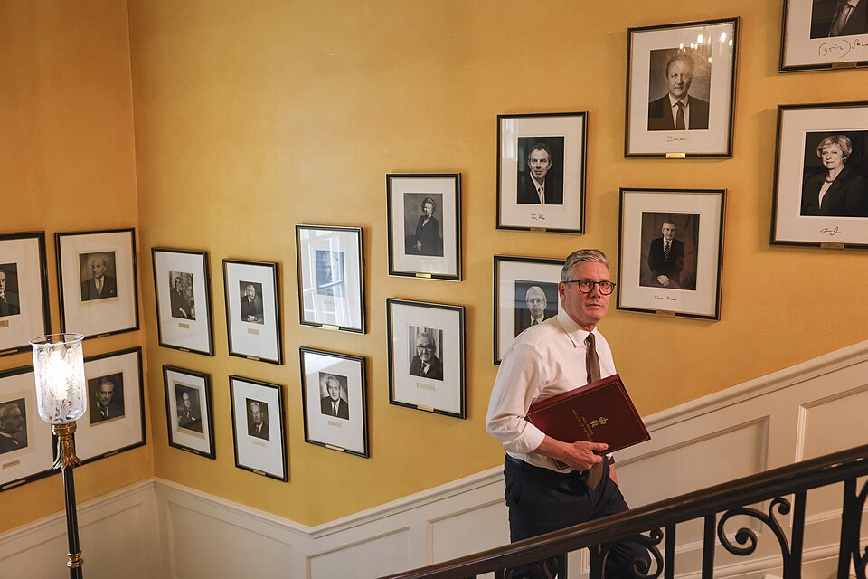Keir Starmer, a middle-aged white man, walks up a flight of stairs past an array of photogarphs of British former prime ministers.