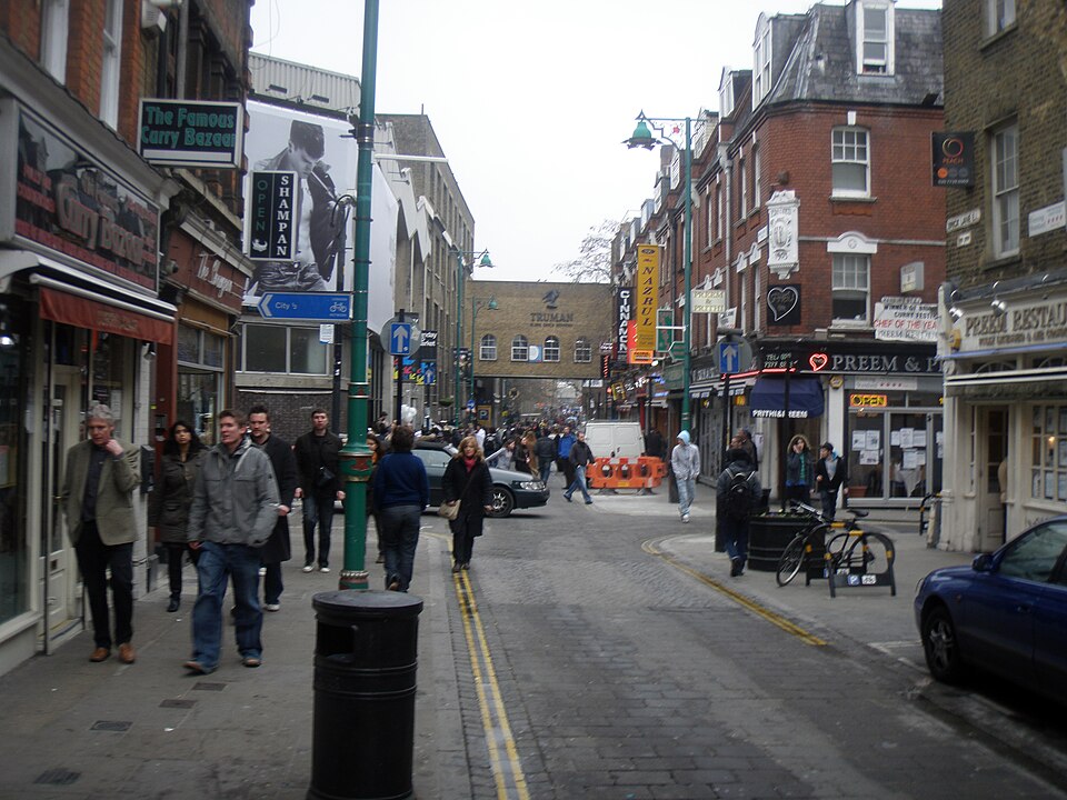 The view up a busy street toward an old yellow brick building with a bridge across labeled Truman Brewery.
